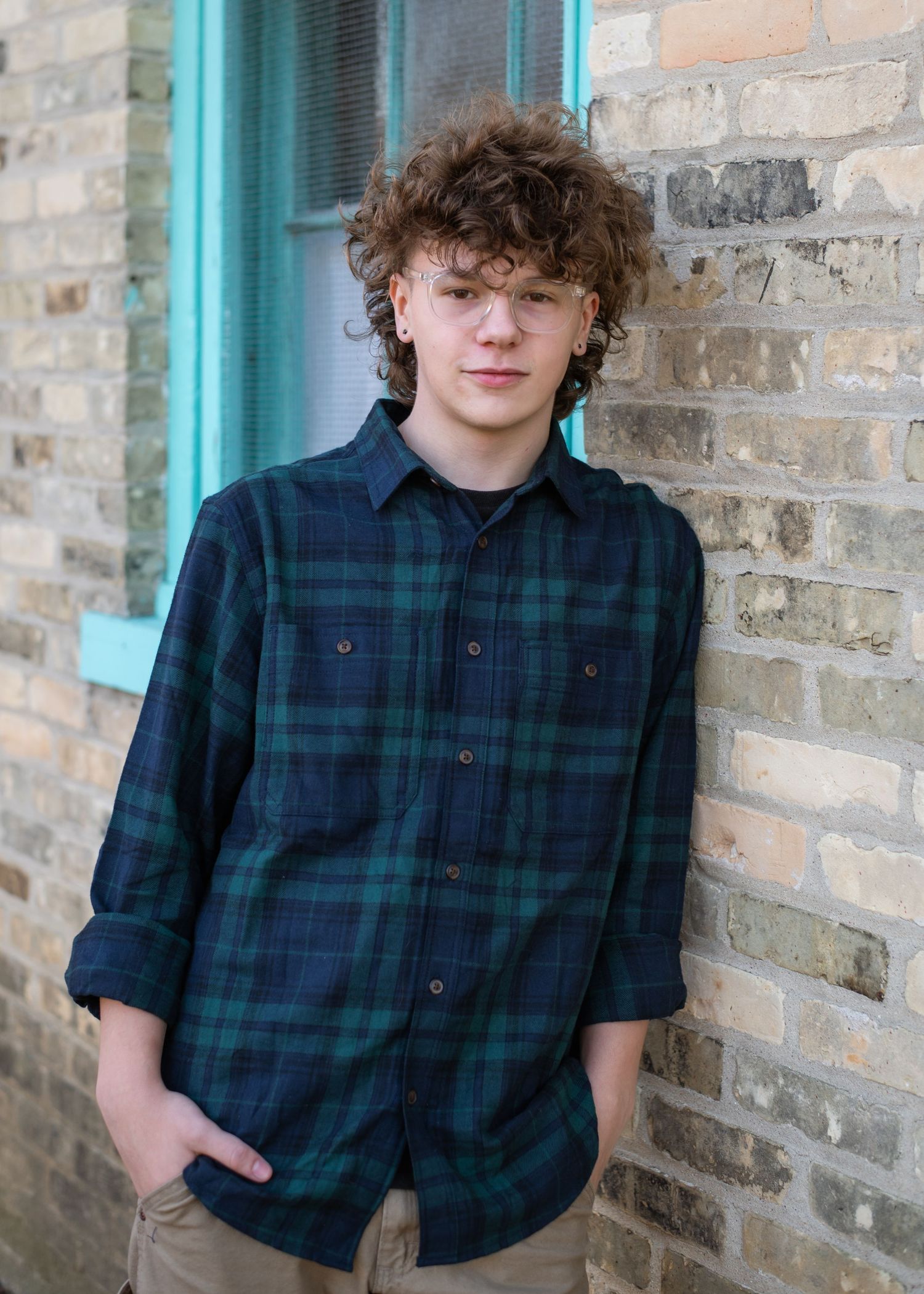 A person in a navy and green plaid button-up shirt poses against a brick wall with turquoise trim.