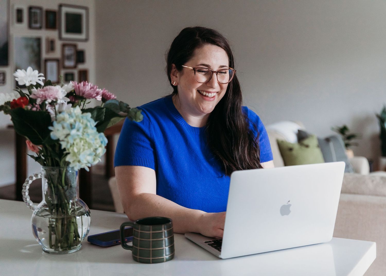 Person in bright blue shirt works on laptop at white desk with floral arrangement in glass vase.