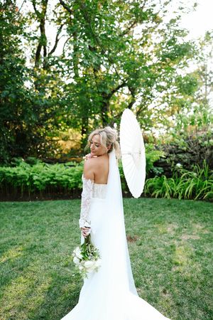 A bride in a white lace dress holds a bouquet while standing in a garden setting with lush greenery.