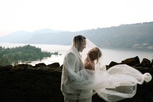 Couple embracing on rocky cliff with flowing veil against mountain lake backdrop.