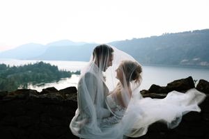 Wedding couple in white attire sharing intimate moment with veil flowing in mountain landscape.