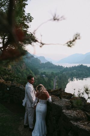 Newlyweds share intimate moment on cliff edge with stunning mountain lake vista.