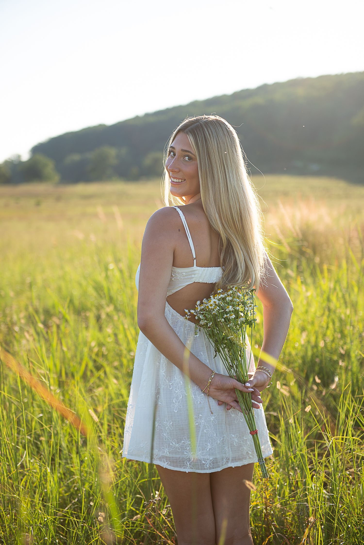 Woman in white summer dress gathers wildflowers in a golden sunlit meadow at sunset.