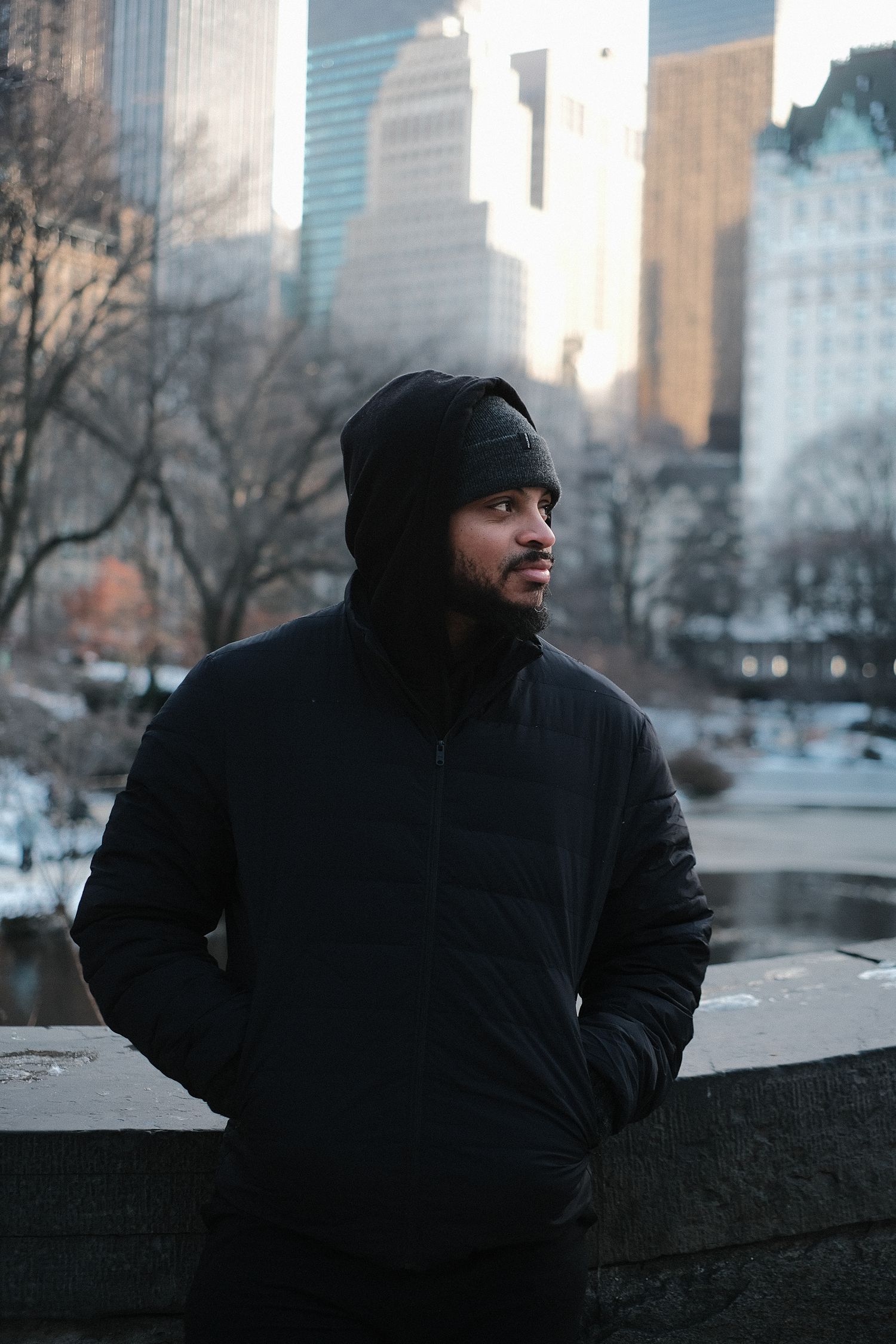 Person in dark hoodie sitting against stone wall with urban skyline backdrop.