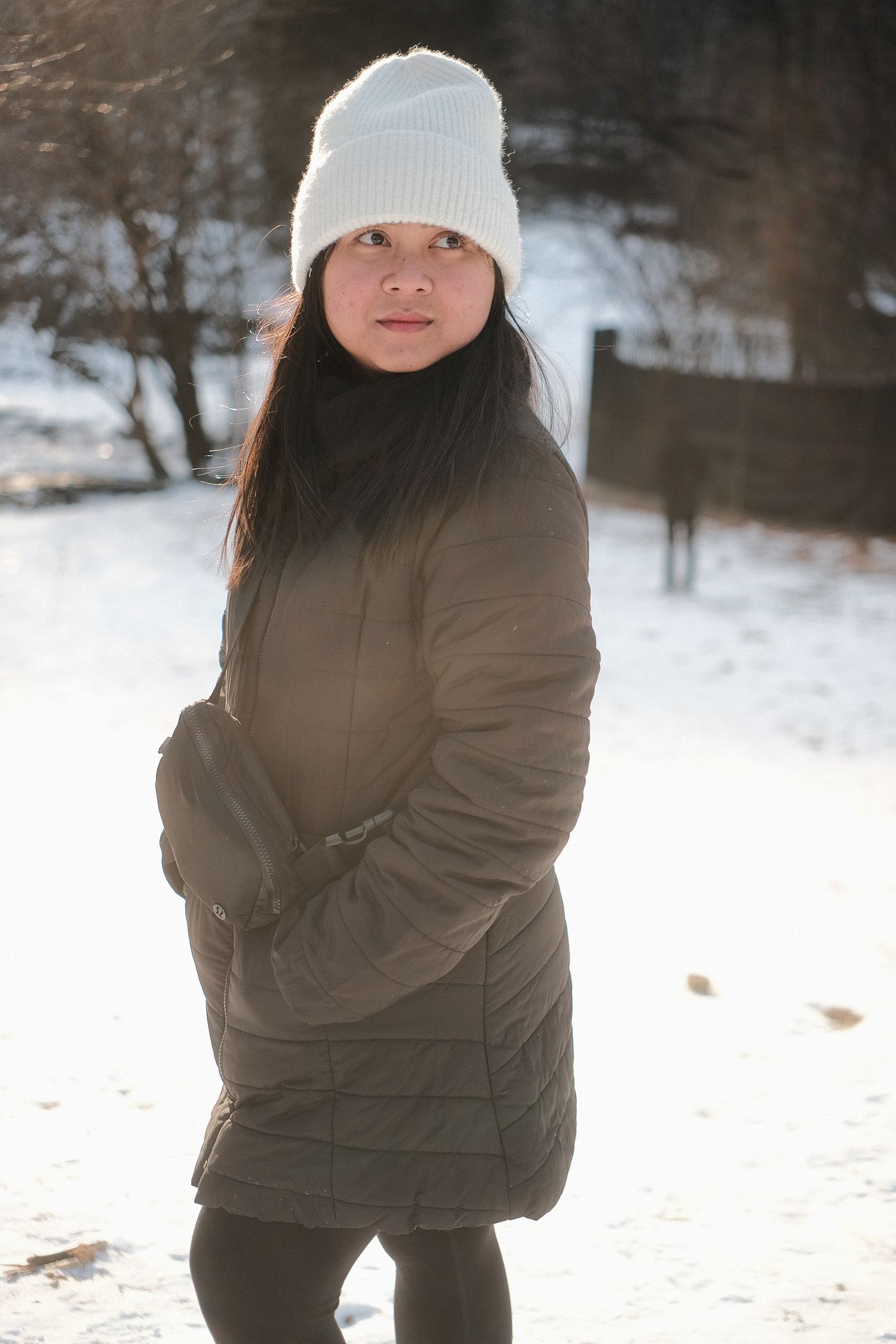 Person wearing brown winter coat and white beanie standing in snowy landscape.
