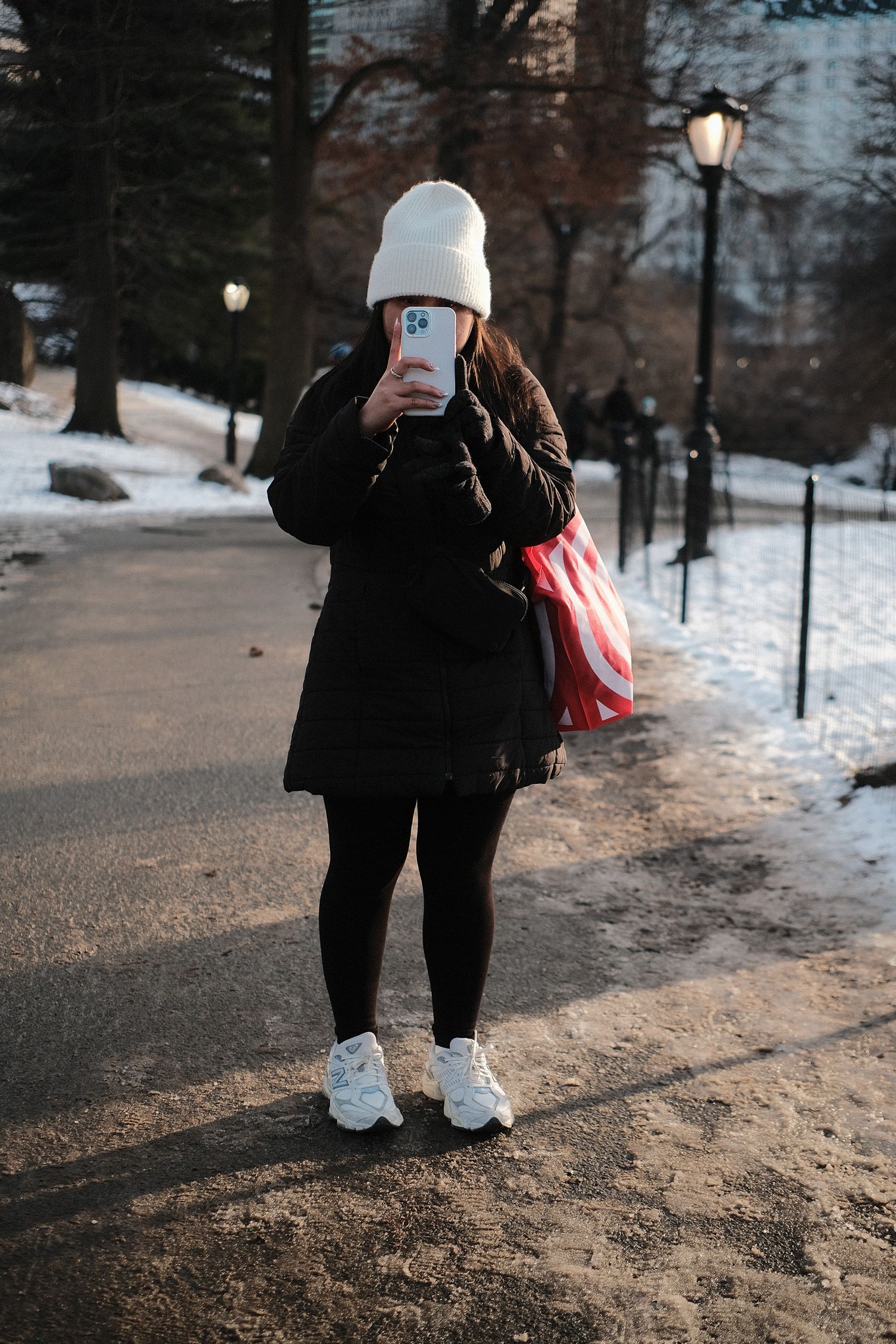 Person in winter clothing standing on a snowy path with lampposts.