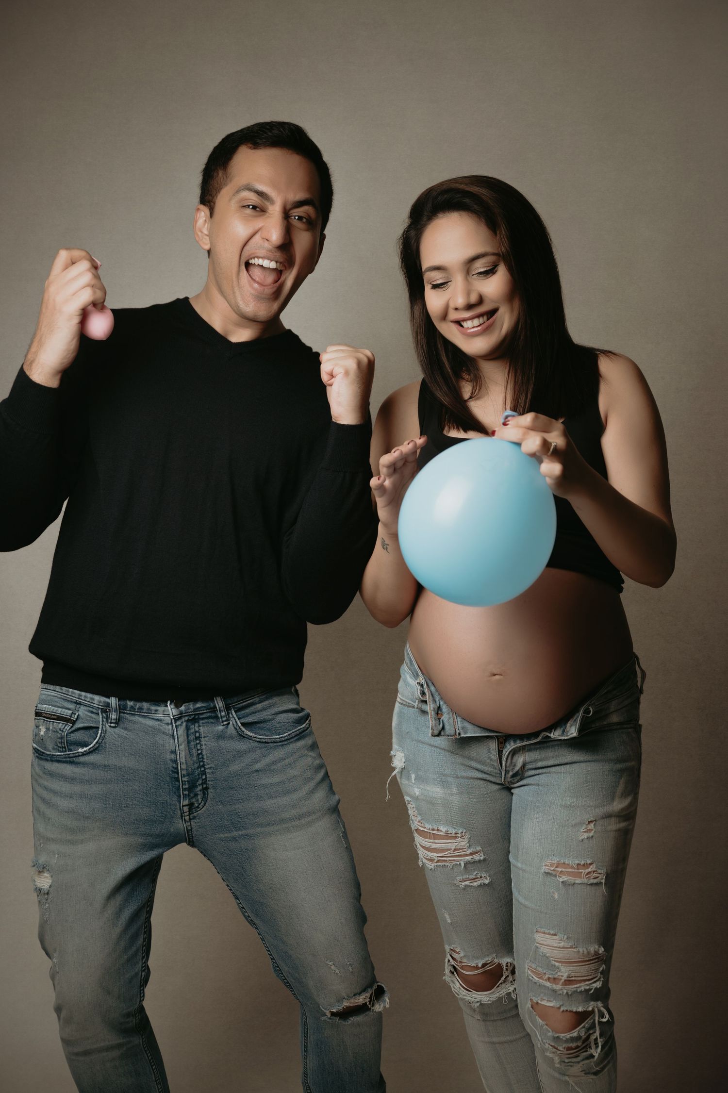 A couple in black tops and ripped jeans poses playfully with a blue balloon in a progressive pregnancy announcement series.