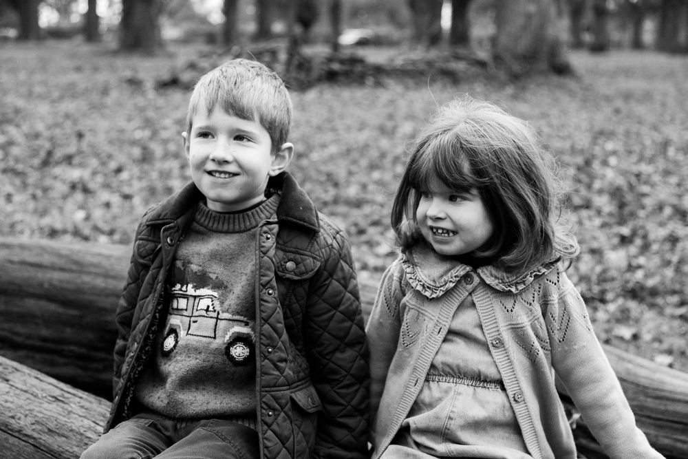 Two children sit together on a wooden log in an autumn forest surrounded by fallen leaves.