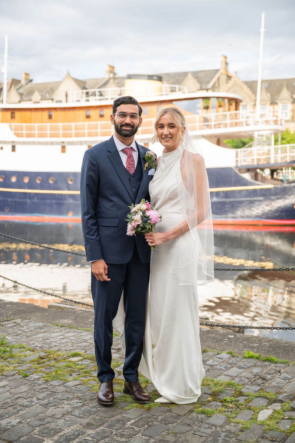 Wedding couple posing in front of a docked boat wearing elegant attire.