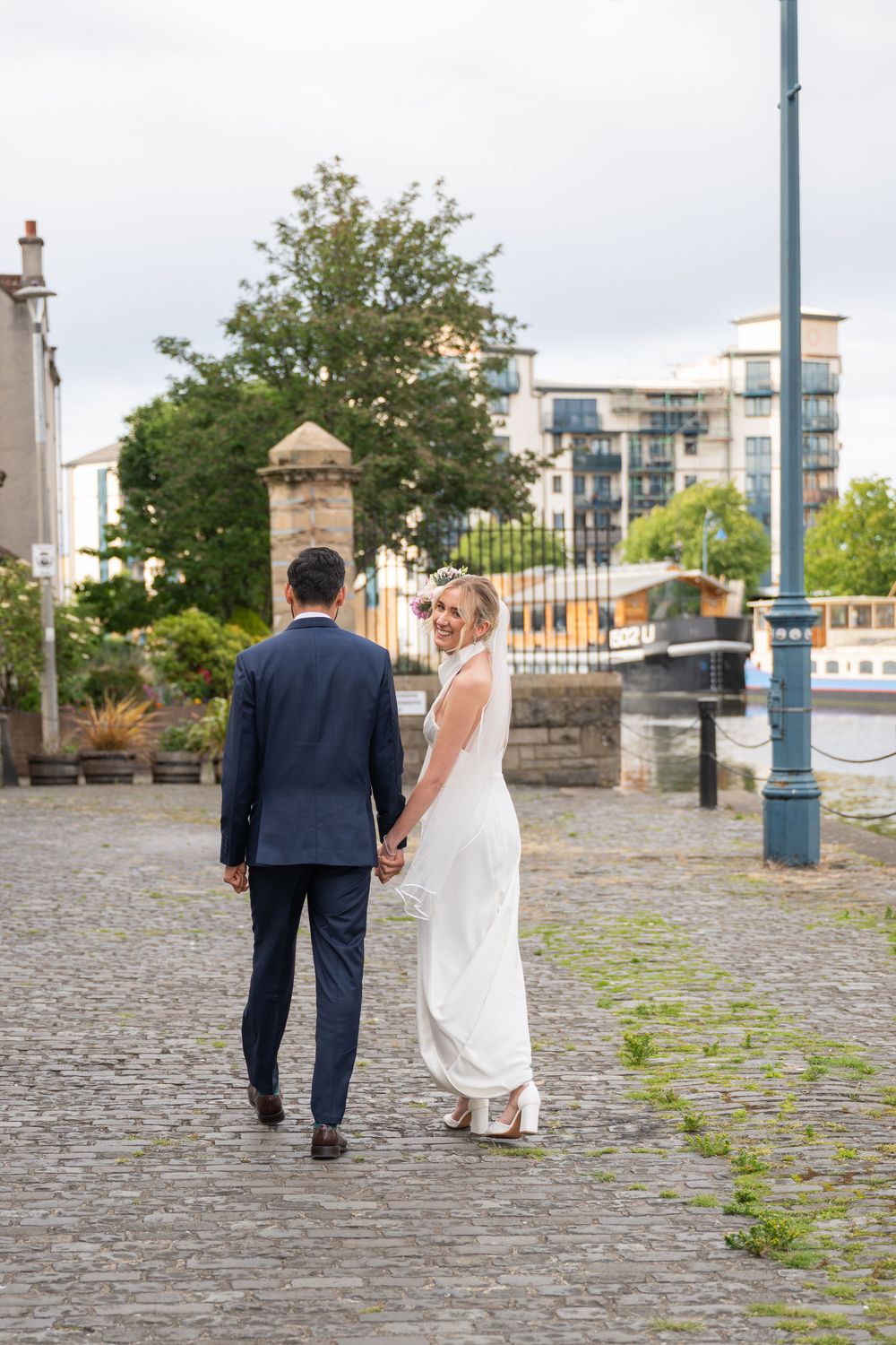 Couple holding hands while walking on cobblestone path near waterfront.