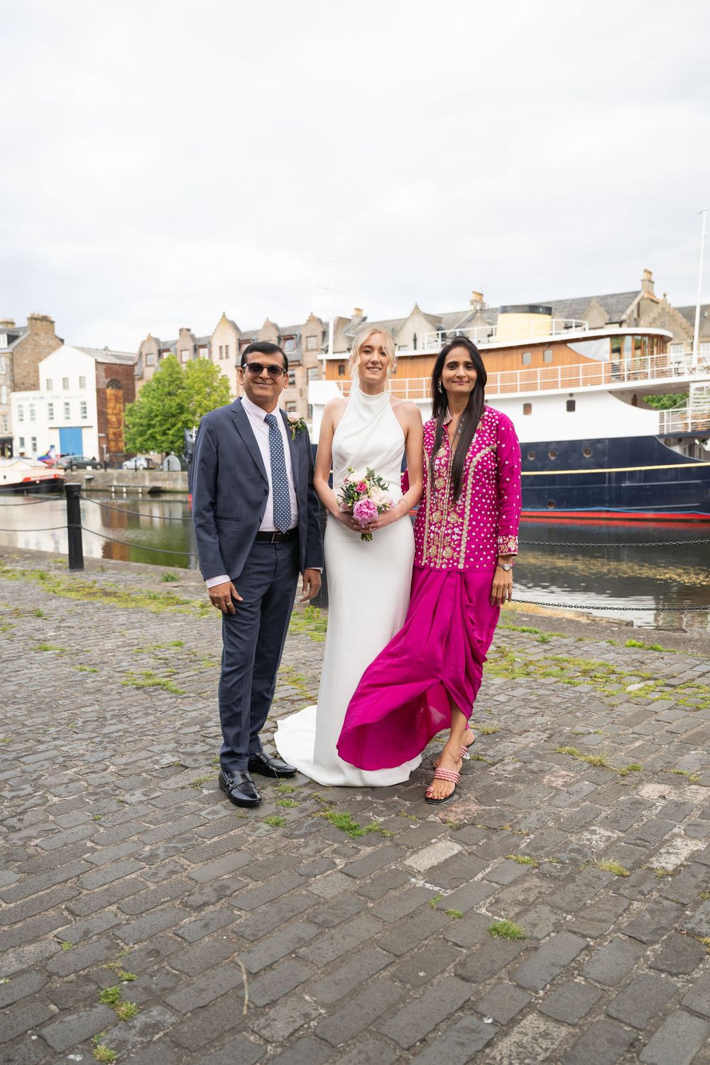 Wedding party posing by harbor with boat and historic buildings in background.