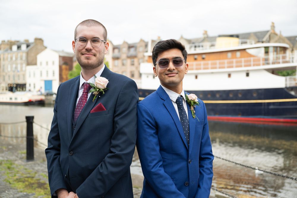 Two wedding party members in navy and royal blue suits pose together by a waterfront.