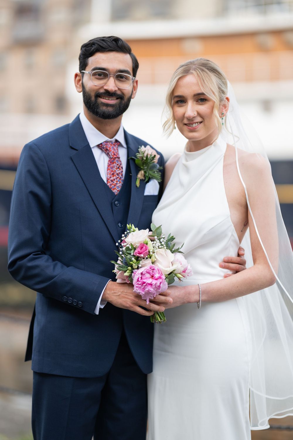 A bride in white dress holds pink bouquet while posing with groom in navy suit.