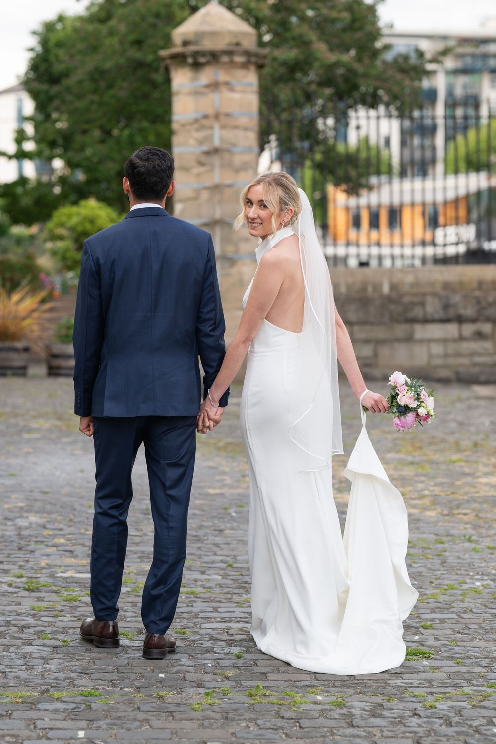 A couple walks hand in hand on cobblestone path with bride looking back over her shoulder.