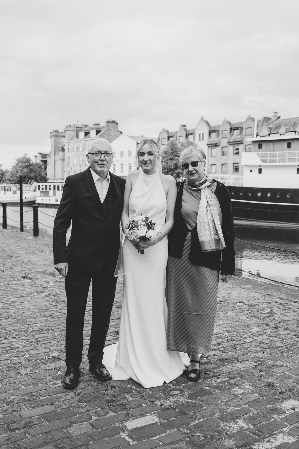 Wedding portrait by the waterfront with a boat and historic buildings in the background.