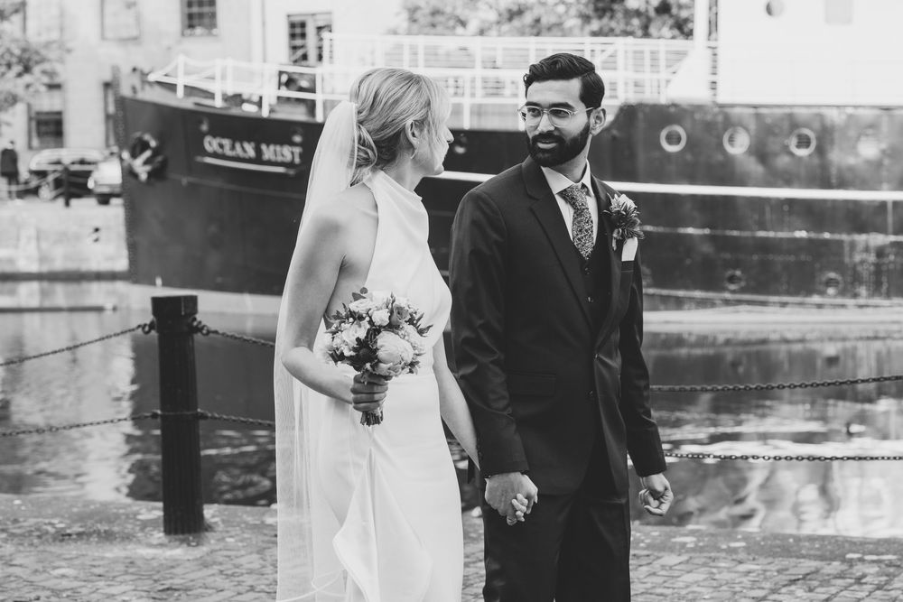 Black and white photo of newlyweds standing by a boat dock holding hands.