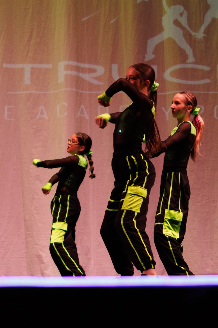 Dancers in black and neon yellow outfits perform a synchronized dance routine on a dimly lit stage.