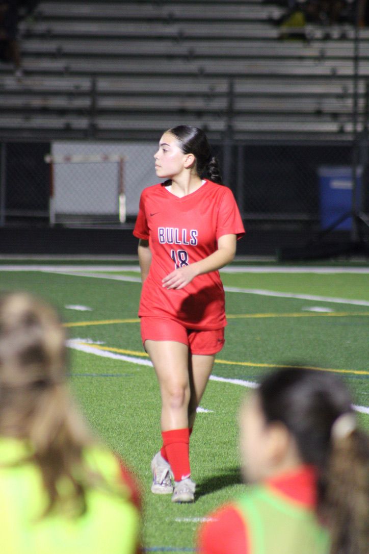 Soccer player in red Bulls uniform walks across field during nighttime game on artificial turf.