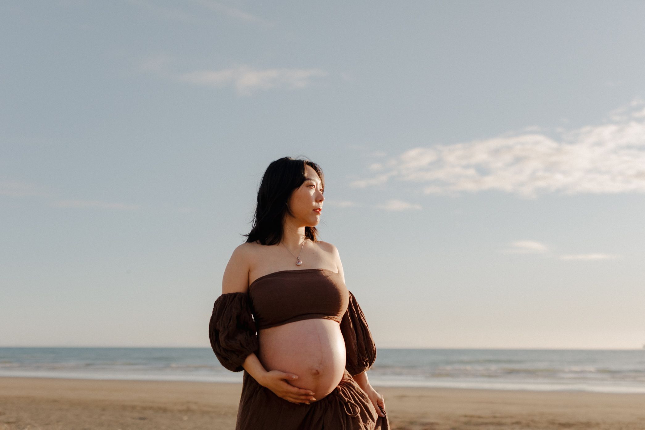 A maternity photoshoot at sunset shows a pregnant woman in a brown dress walking along a serene beach.