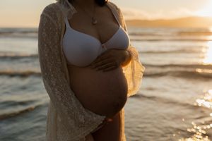 A pregnant figure in a white bikini top and lace cardigan stands on a beach at sunset, cradling their belly.