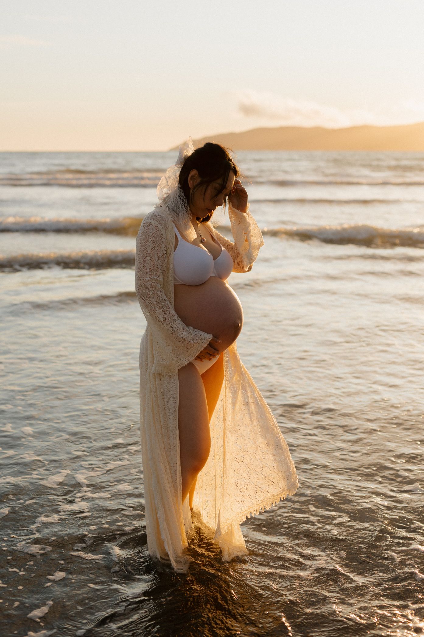 A romantic couple embraces on a beach at sunset with waves lapping at their feet as golden light illuminates their silhouettes.