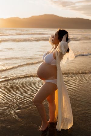 Silhouetted figure in white dress stands at sunset beach edge with gentle waves lapping at shore in golden light.