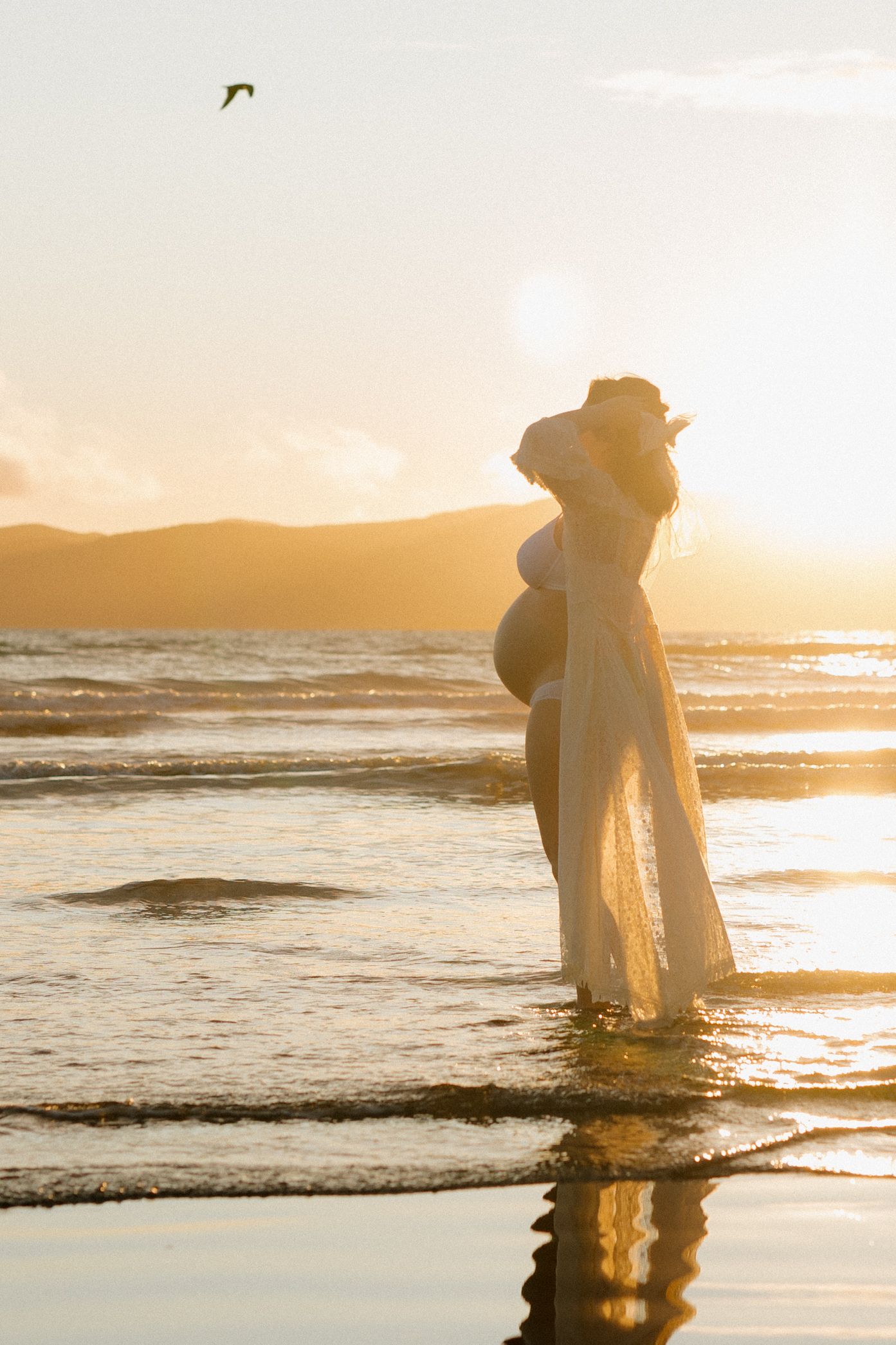 Silhouette of a pregnant figure in a flowing dress standing in ocean waves during golden sunset.