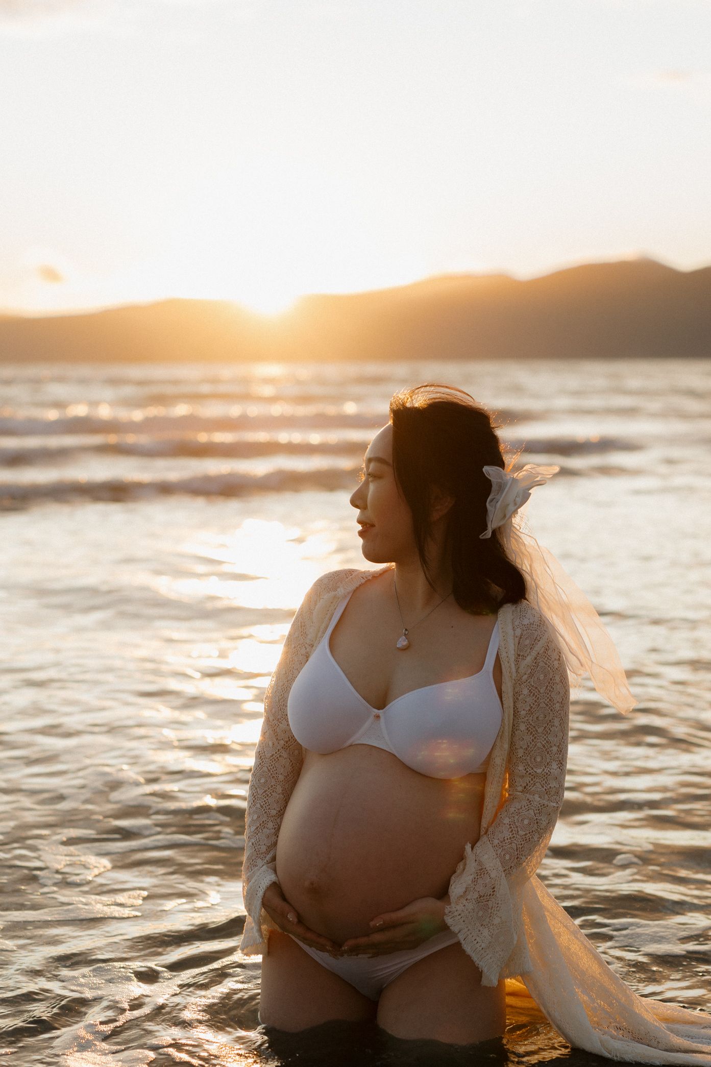 Expectant mother in white attire poses at beach during warm sunset with waves gently lapping at shore.