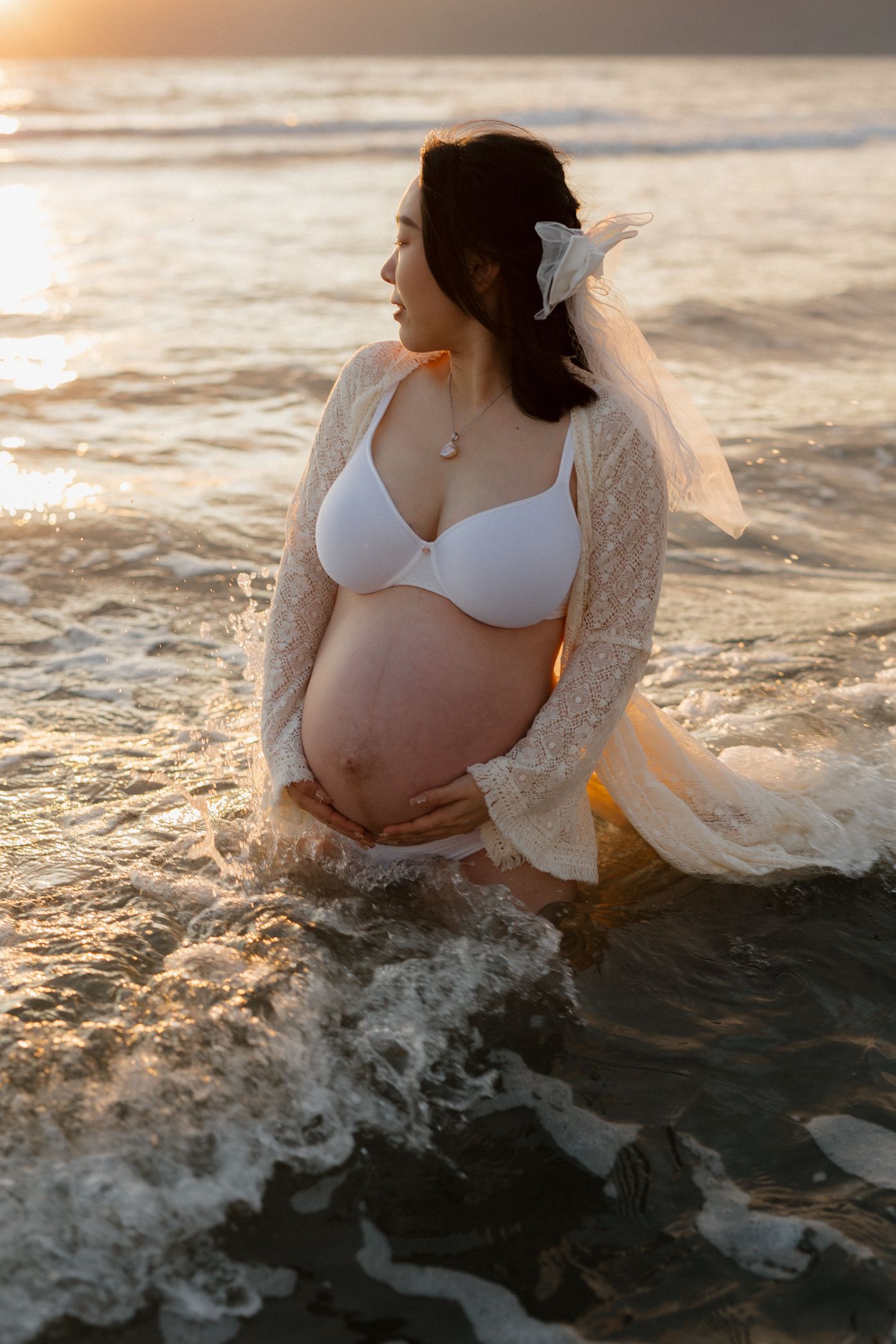 Expectant mother in white lace robe sits in ocean waves during golden hour maternity photoshoot.