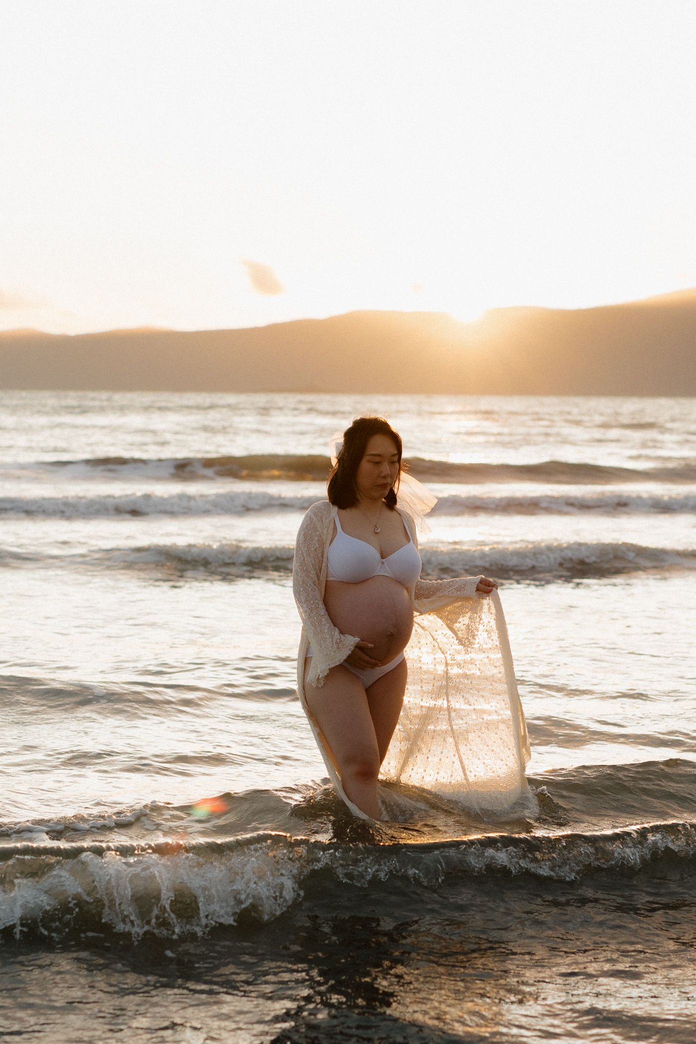 A woman in white swimwear poses gracefully in ocean waves during golden hour sunset.