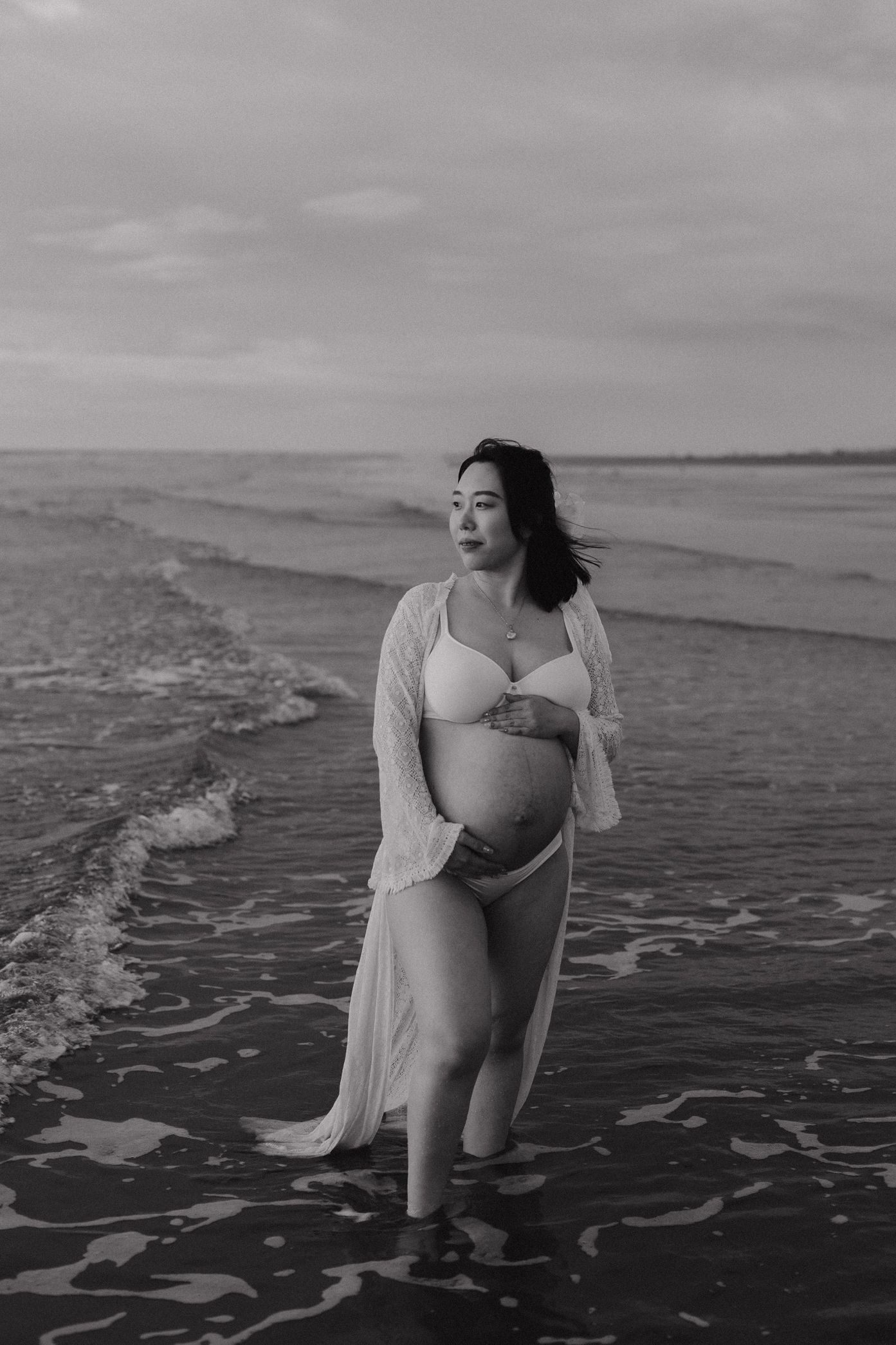 Black and white photo of a pregnant woman in white dress standing in shallow ocean water.