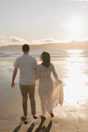 A couple walks hand in hand along a serene beach at sunset, their silhouettes casting long shadows on the sand.