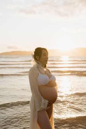 Silhouette of an expectant mother in a white dress standing on a beach at sunset with waves in the background.