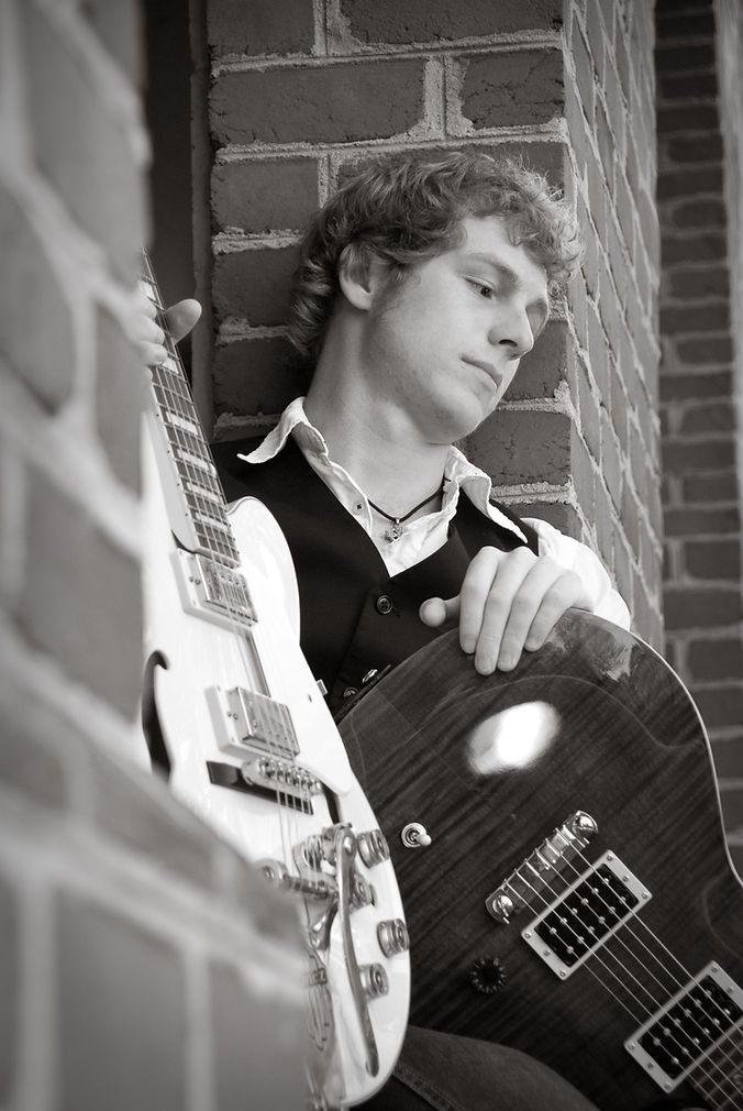 Black and white photo of a young man leaning against a brick wall holding two electric guitars.