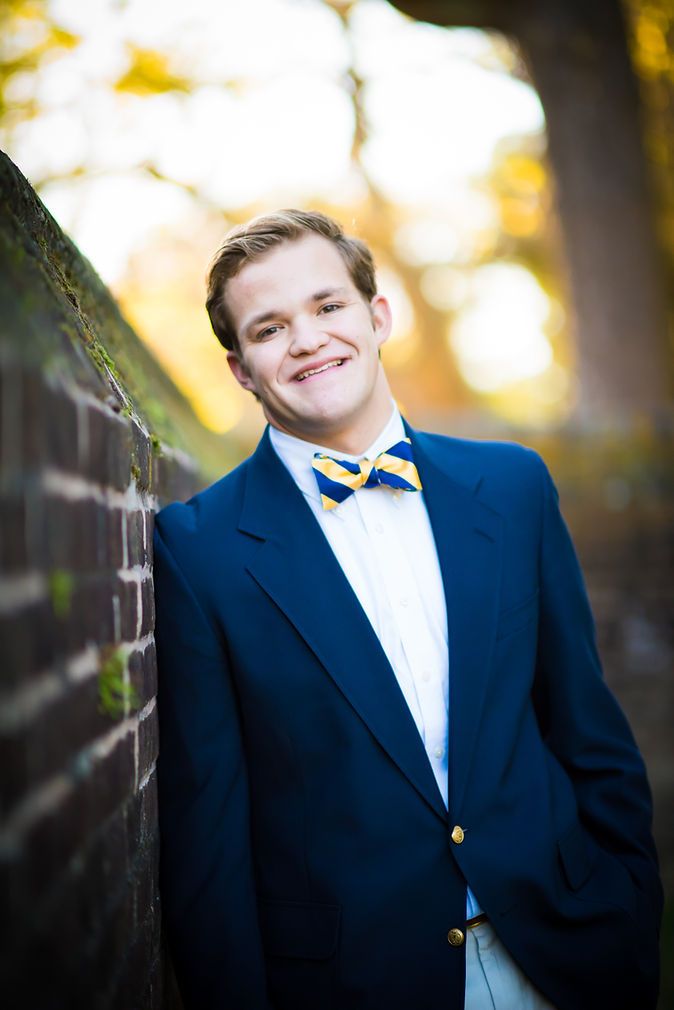 Young man in navy blazer and gold bow tie leaning against brick wall outdoors.