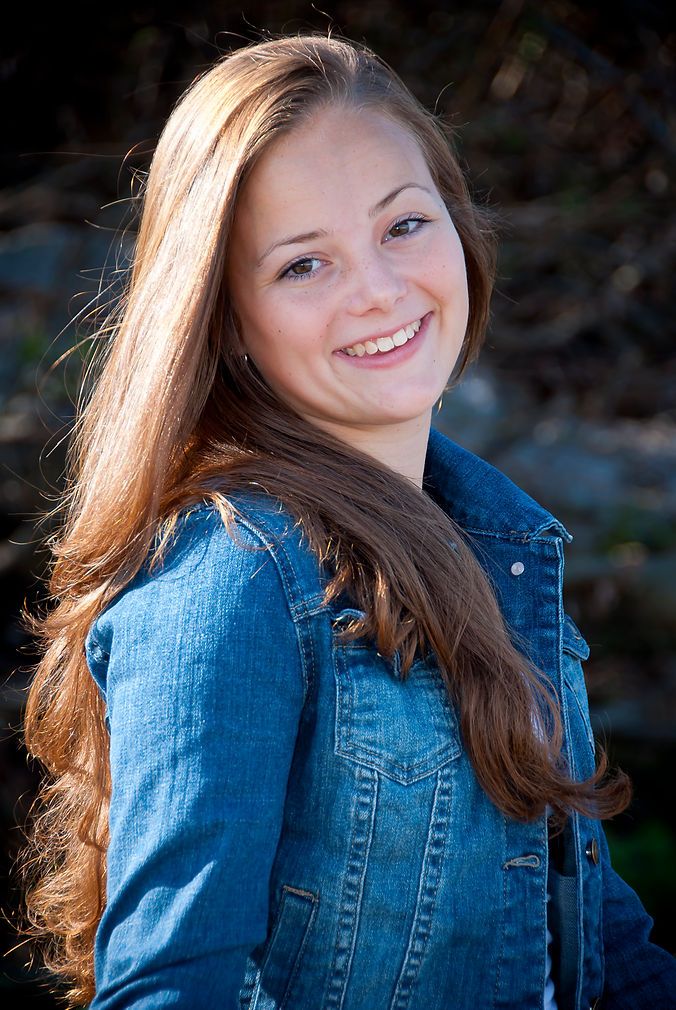 Smiling young woman with long auburn hair wearing a blue denim jacket outdoors in natural sunlight.