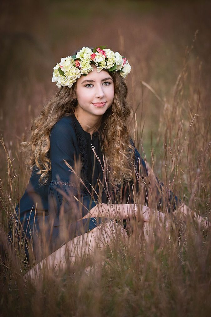Young woman with curly hair wearing a floral crown sitting in a field of tall golden grass.