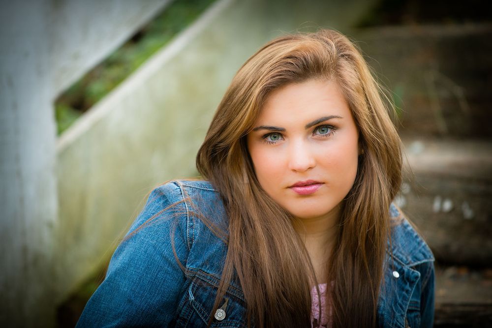 Young woman with long brown hair in a denim jacket sitting on outdoor concrete steps.