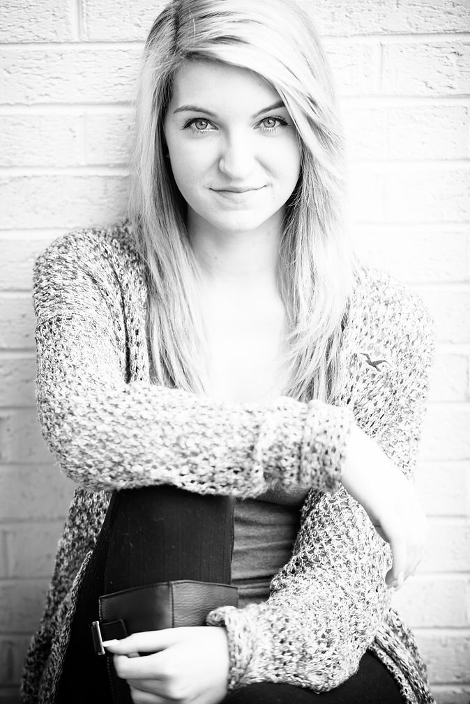 Black and white portrait of a blonde woman in a knit cardigan leaning against a brick wall.