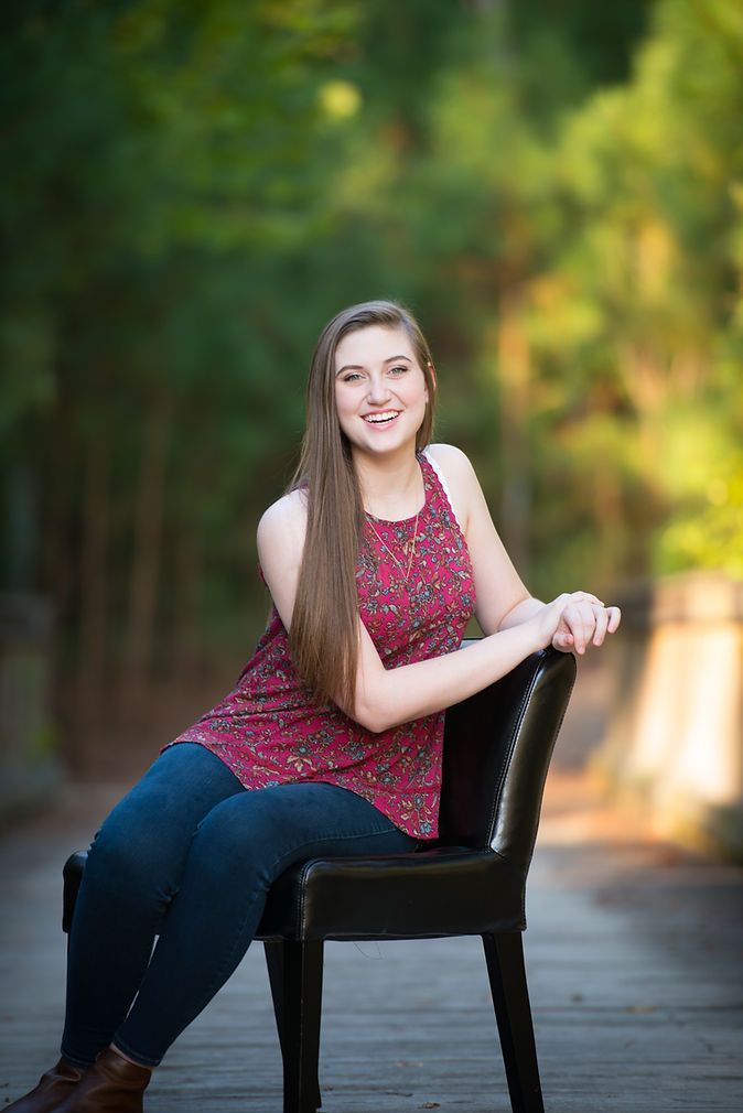 Smiling teen girl in floral top seated on chair outdoors among green trees.
