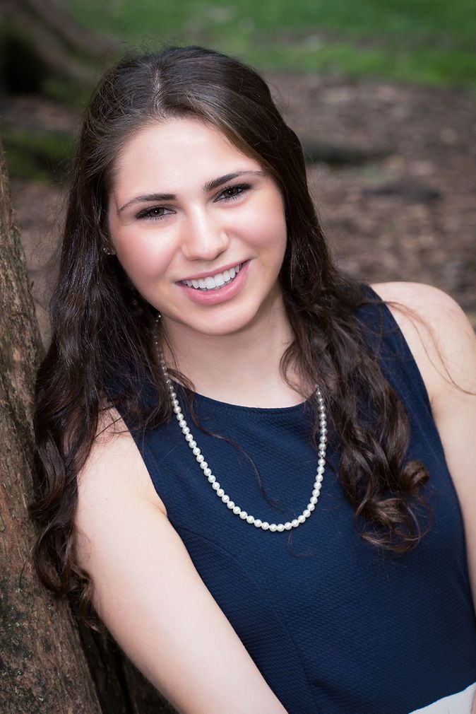 Smiling brunette teen girl in navy dress with pearl necklace leaning against tree.