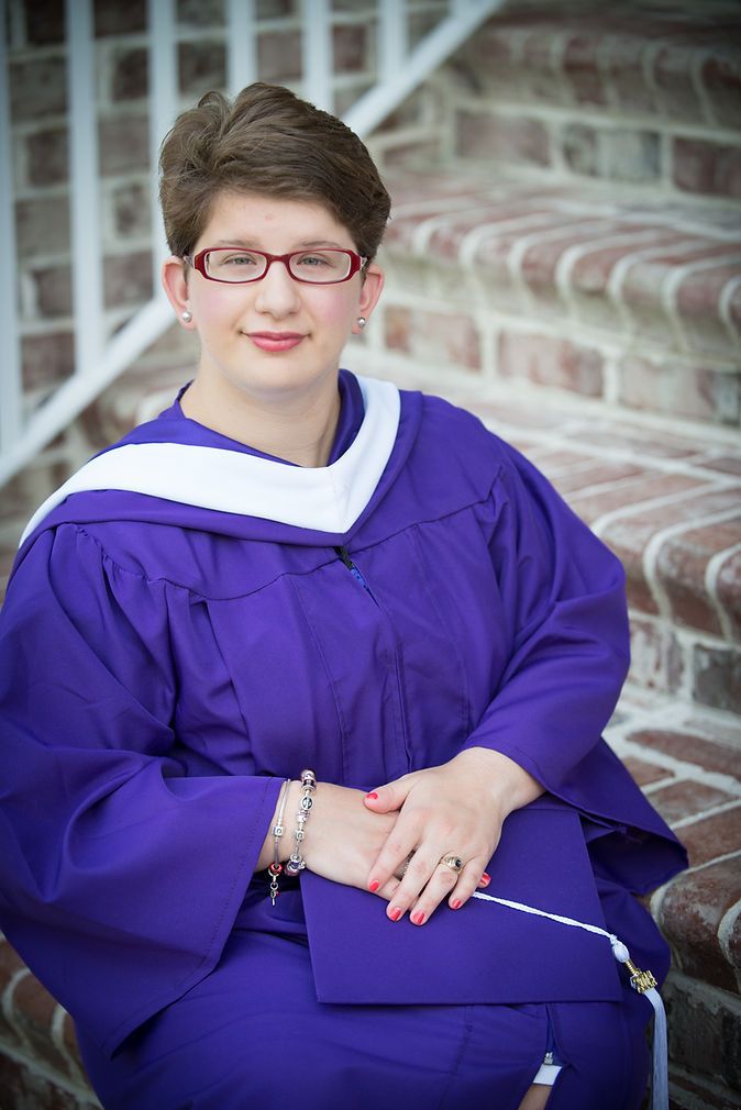 Graduate in purple cap and gown sitting on brick steps holding diploma during graduation ceremony.