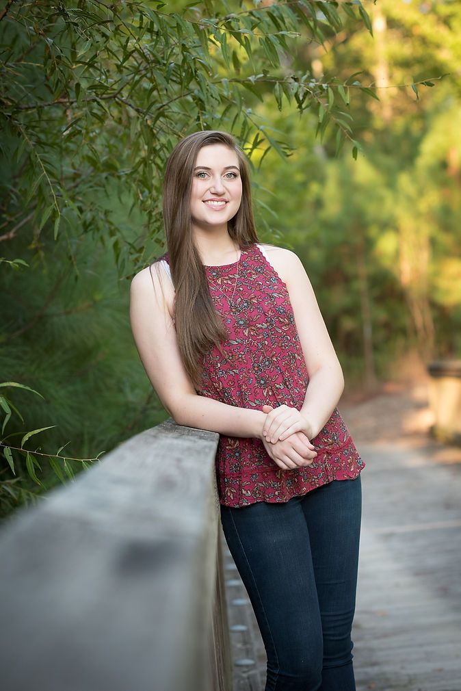 Smiling teen girl in floral top leaning on wooden bridge railing surrounded by greenery.