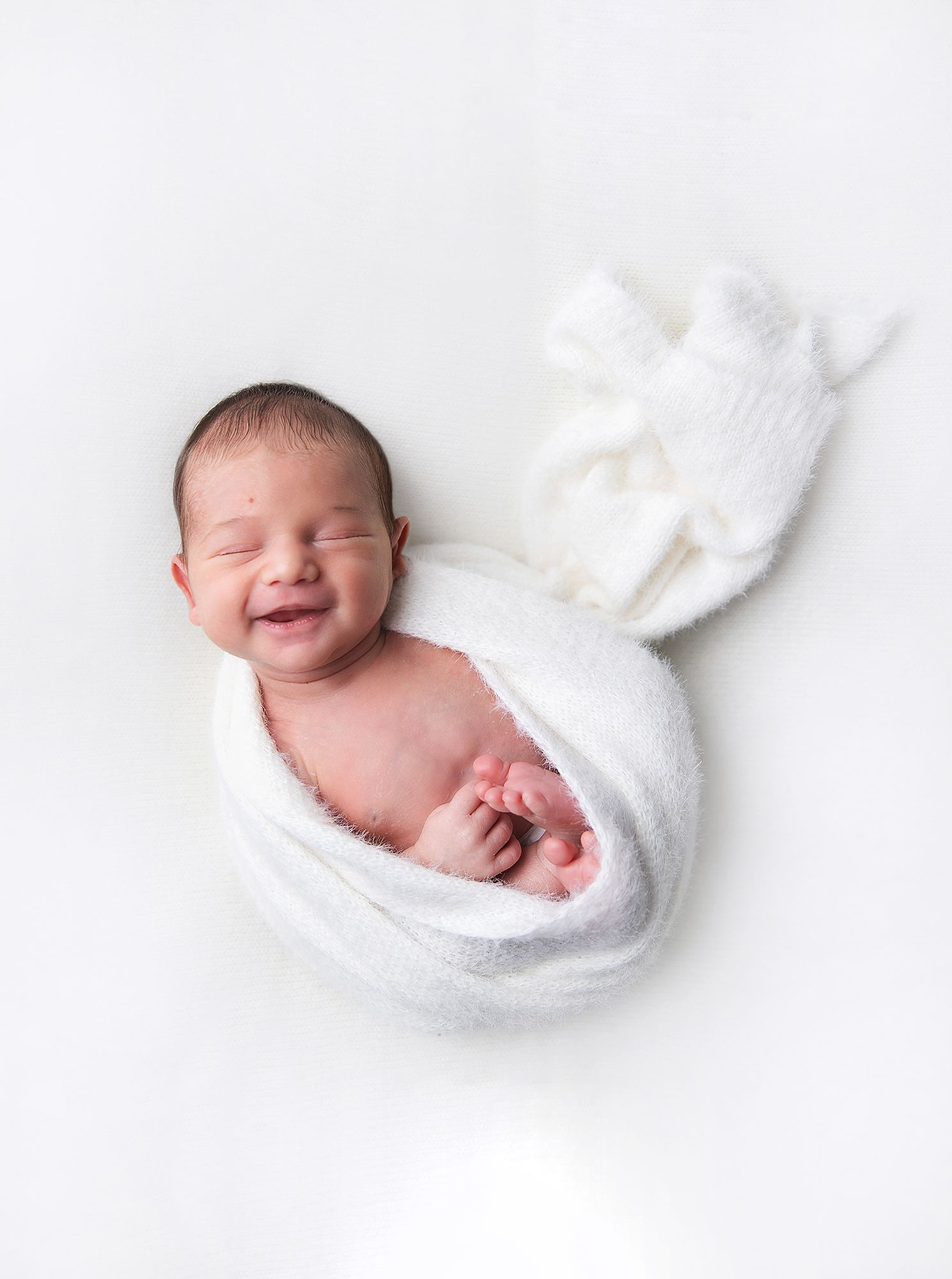 A sleeping newborn baby wrapped in soft white blankets against a white background. During a newborn photoshoot in Bexley