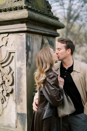 A romantic couple shares a tender kiss while standing near an ornate stone pillar in a park setting.