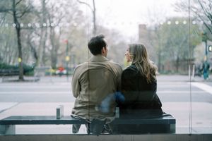 Two people sit together on a bench in an urban setting as seen through multiple glass window reflections.