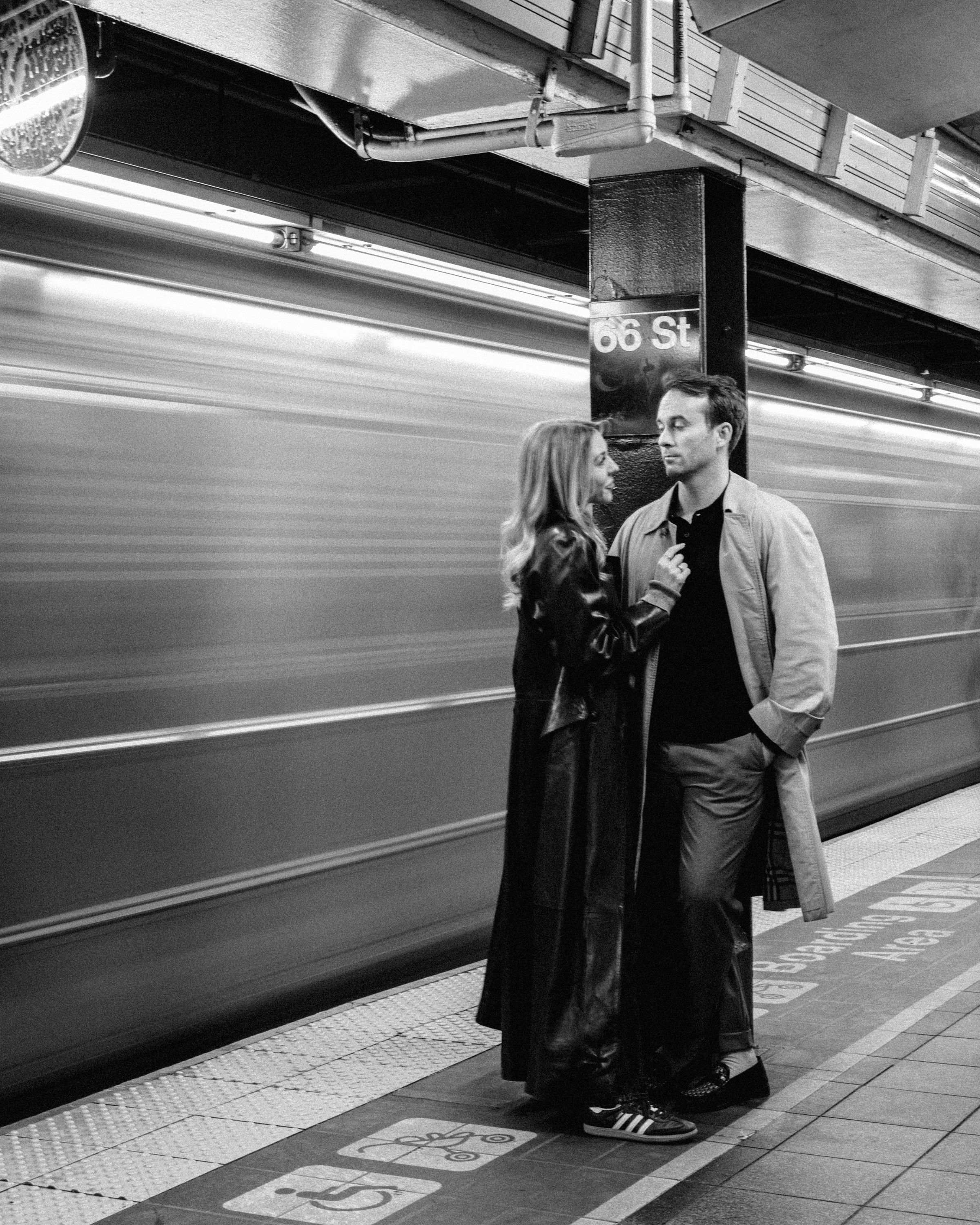 A couple stands on a subway platform as a blurred train speeds past in black and white.