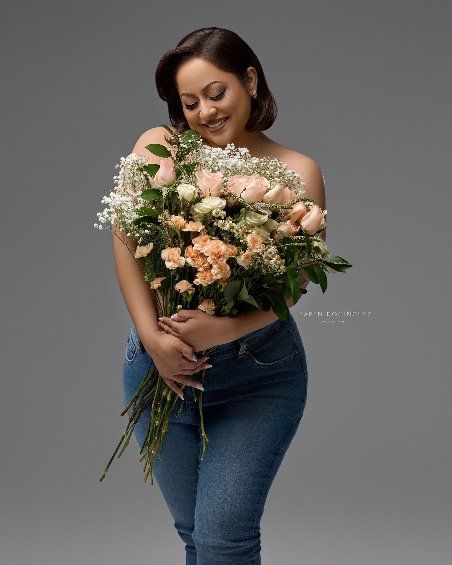A person in jeans holds a lush bouquet of peach roses and baby's breath against a gray studio background.