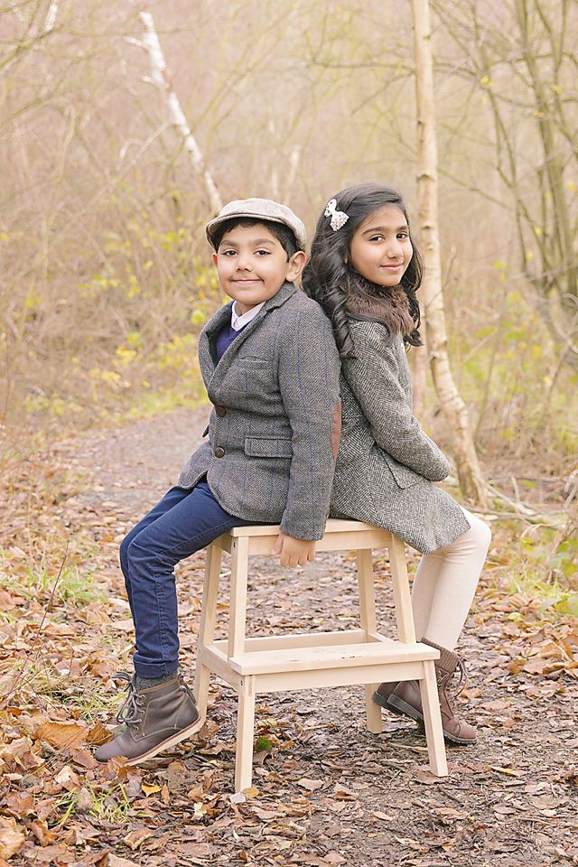 Two children sit back-to-back on a wooden bench surrounded by fallen autumn leaves in a forest setting.