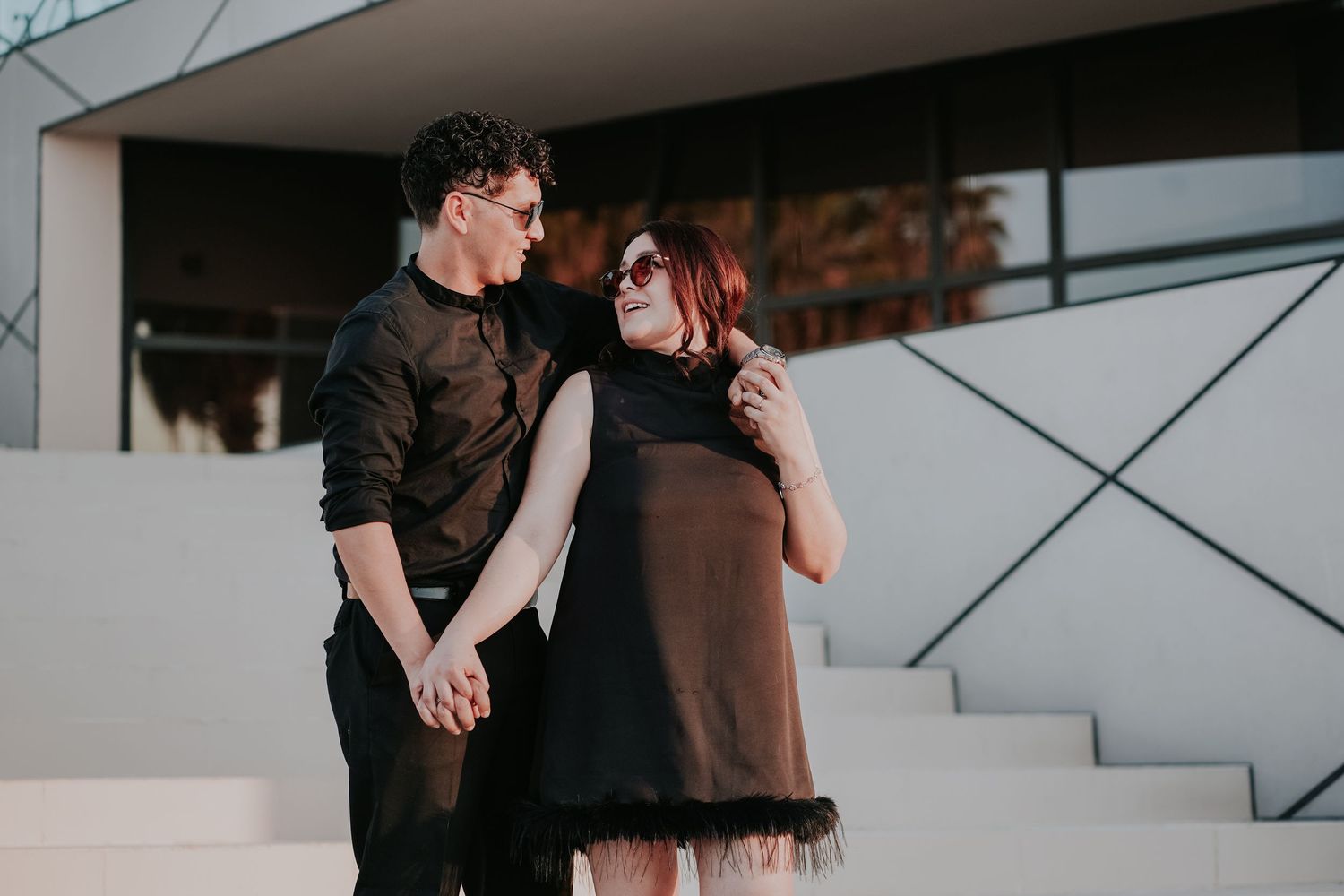 Two people in dark clothing share a romantic moment on minimalist white stairs.